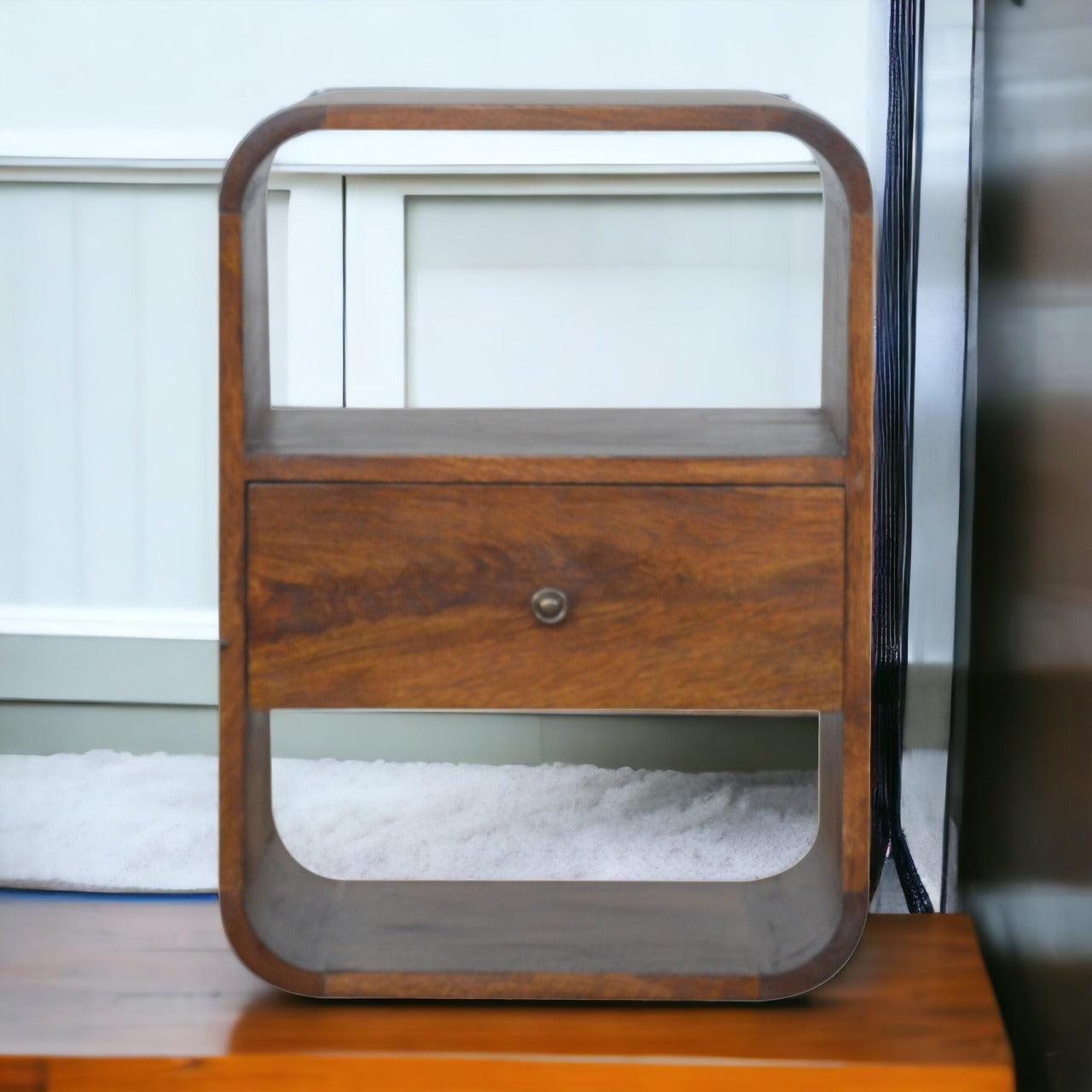 Chestnut Curved Edge Nightstand made from solid mango wood with drawer and two open storage slots. Nightstand sitting on a bench.